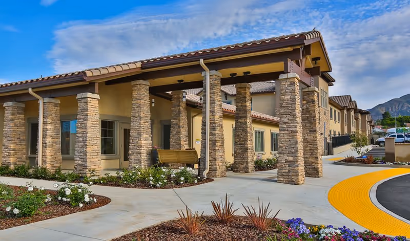 Exterior view of Brightwater Senior Living of Highland showing a building with stone pillars, beige walls, and a tiled roof under a partly cloudy sky. There are landscaped flower beds with white and purple flowers along a curved sidewalk with a yellow tactile paving strip.