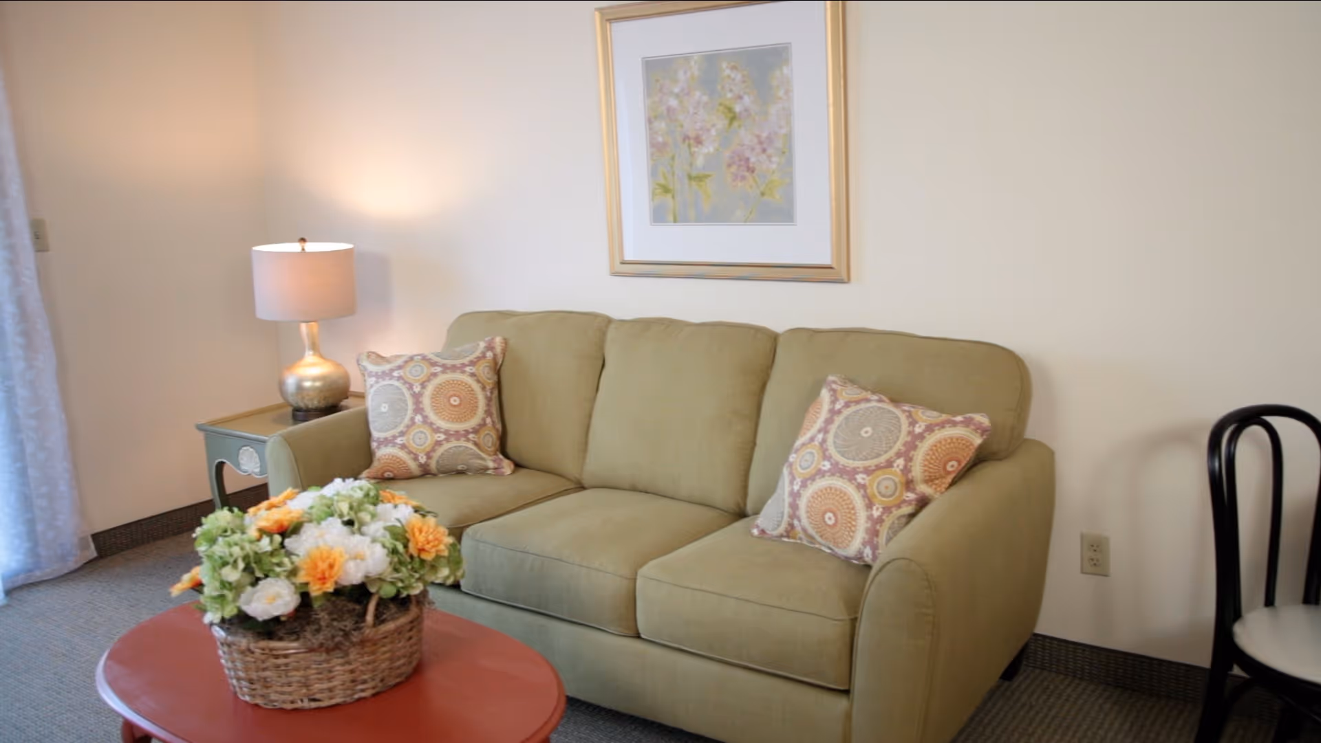 A cozy living room area with a light green sofa adorned with two patterned throw pillows. A small red coffee table in front holds a basket of colorful flowers. To the left, there is a side table with a lamp, and a framed floral artwork hangs on the wall above the sofa. A black chair with a white cushion is partially visible on the right.