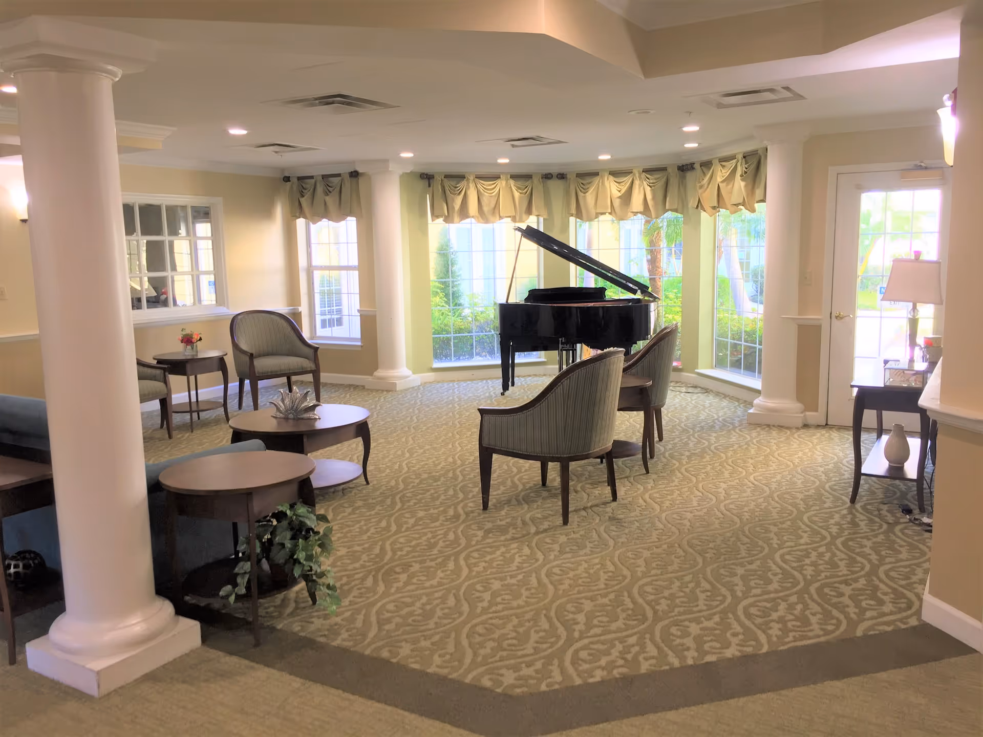 A bright and spacious living room area with large windows letting in natural light. The room features a black grand piano near the windows, several upholstered chairs, round wooden tables, decorative columns, and patterned carpet. There is a door leading outside and a lamp on a side table.