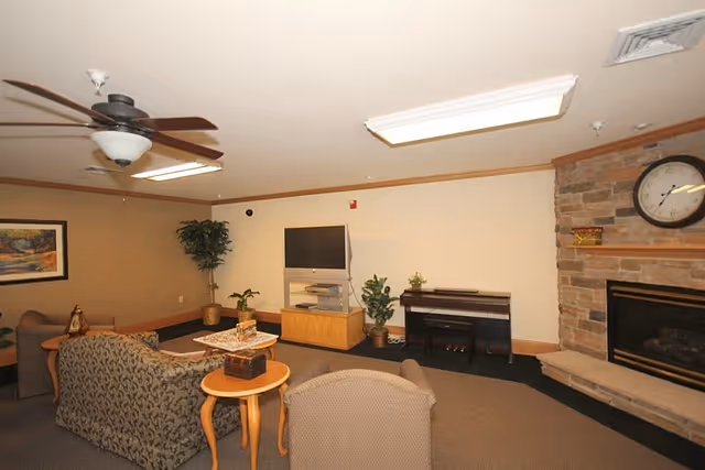 Cozy senior living lounge with sofas, a television, plants, and a stone fireplace.