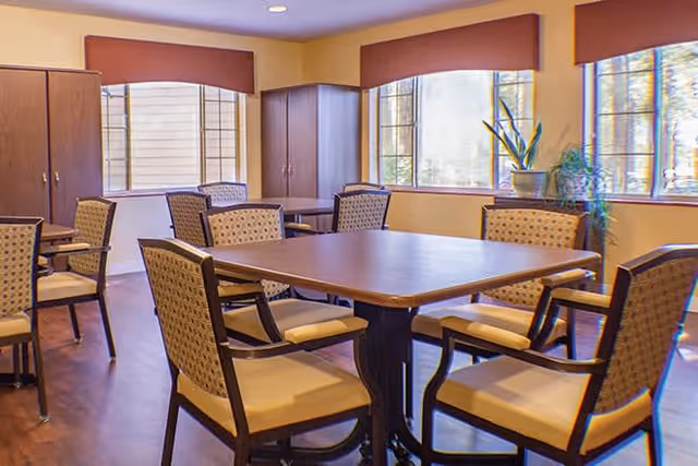 A bright dining room with a large square wooden table surrounded by eight cushioned chairs with patterned backs. The room has large windows with brown valances, letting in natural light, and wooden cabinets along the walls. A tall plant is placed near the windows.