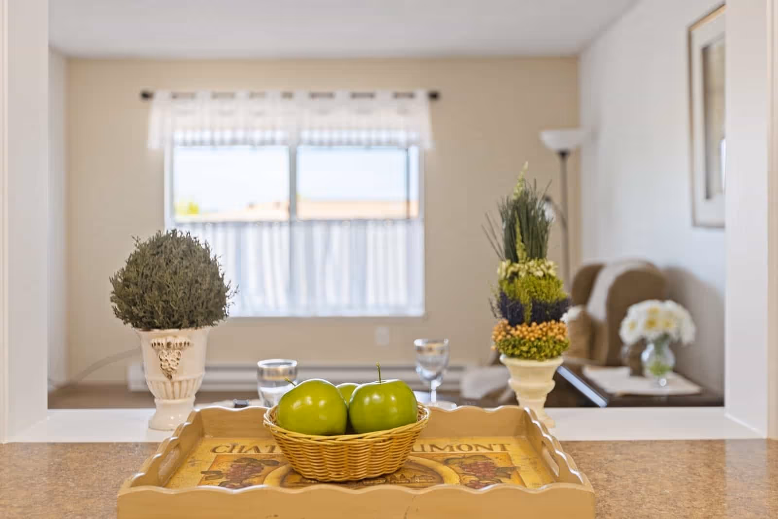 A cozy living room area viewed through a pass-through window from the kitchen. On the counter in the foreground is a decorative tray holding a small basket with three green apples. On either side of the tray are two ornamental plants. In the background, there is a window with sheer white curtains, a beige armchair, a floor lamp, and a side table with a vase of white flowers.