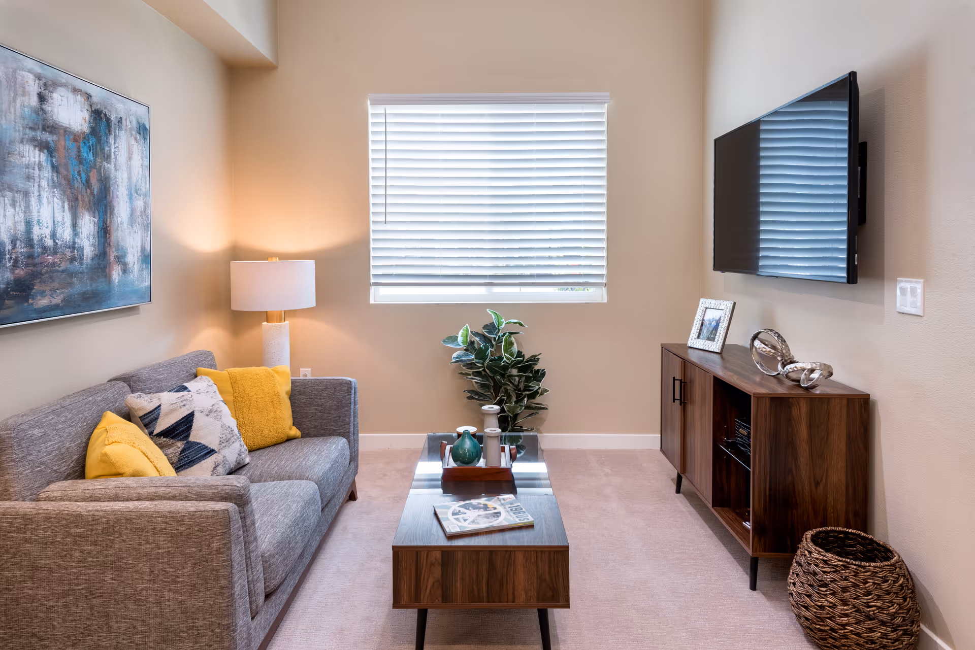A cozy living room with a gray sofa adorned with yellow and patterned pillows, a wooden coffee table with decorative items and a book, a potted plant near a window with white blinds, a wooden TV stand with decorative objects and a framed photo, and a flat-screen TV mounted on the wall.