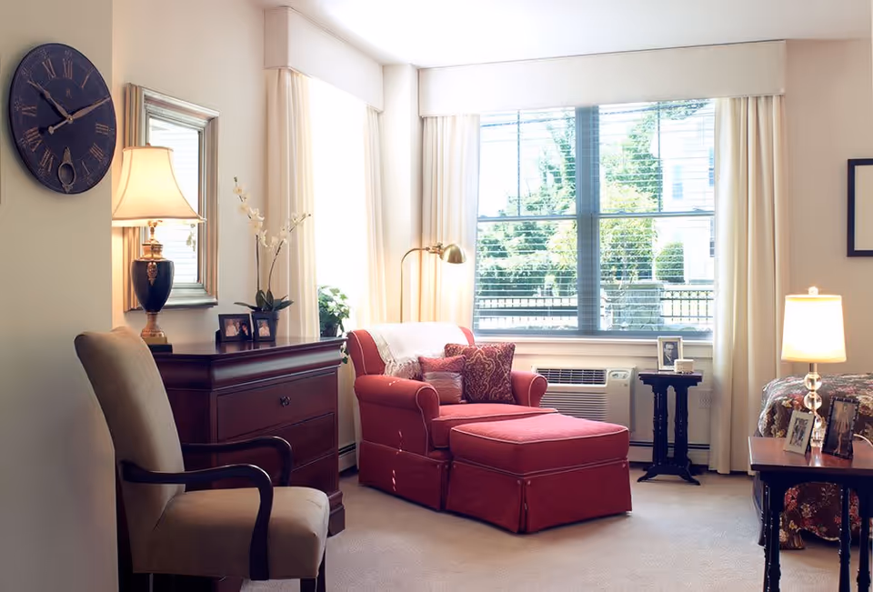 A cozy living area with a large window letting in natural light. The room features a red upholstered armchair with an ottoman, a wooden dresser with a lamp and framed photos, a beige upholstered chair, and a small side table with a lamp and more framed photos. Light-colored curtains frame the window, and the carpet is beige.