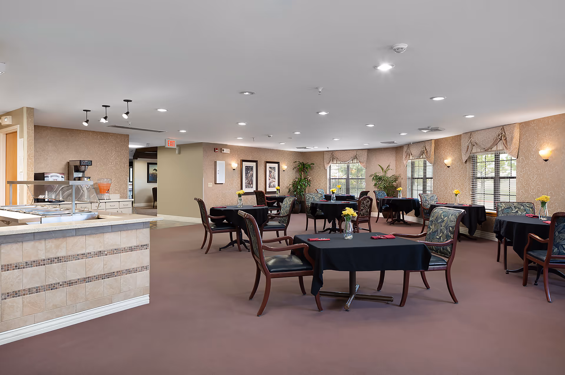 A spacious dining room with multiple tables covered in black tablecloths, each set with a small vase of yellow flowers and red napkins. The room has large windows with decorative valances, patterned wallpaper, and several framed pictures on the walls. There is a serving counter on the left side with a beverage dispenser and trays. The floor is carpeted in a muted brown color, and the ceiling has recessed lighting.