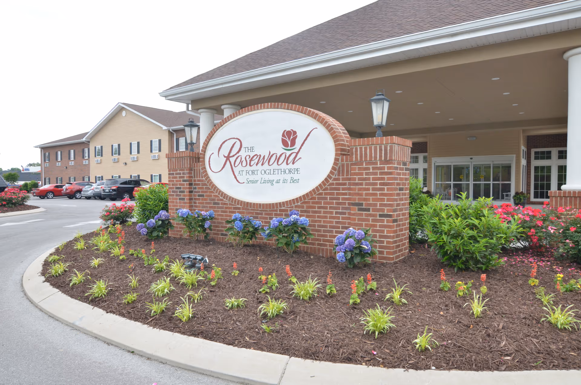 Entrance area of The Rosewood at Fort Oglethorpe senior living facility featuring a large brick sign with the facility's name surrounded by landscaped flower beds with purple and red flowers, and the building with parked cars in the background.