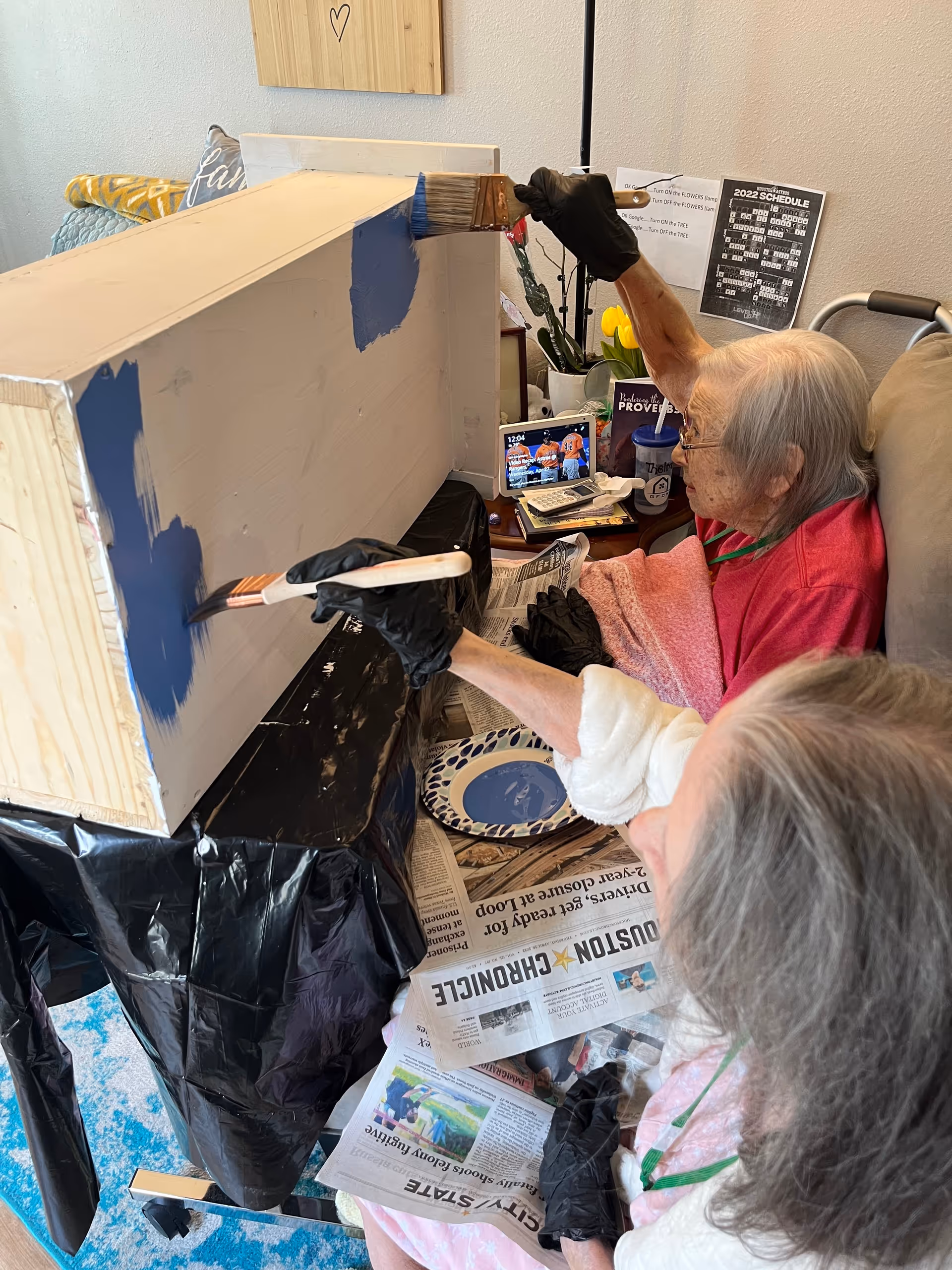 Two elderly women wearing gloves are painting a wooden box with blue paint. They are seated in a cozy room with a small table holding a tablet, a cup, and some papers. One woman is wearing a red top and glasses, while the other is wearing a white top. The room has a calendar and some decorations on the wall.