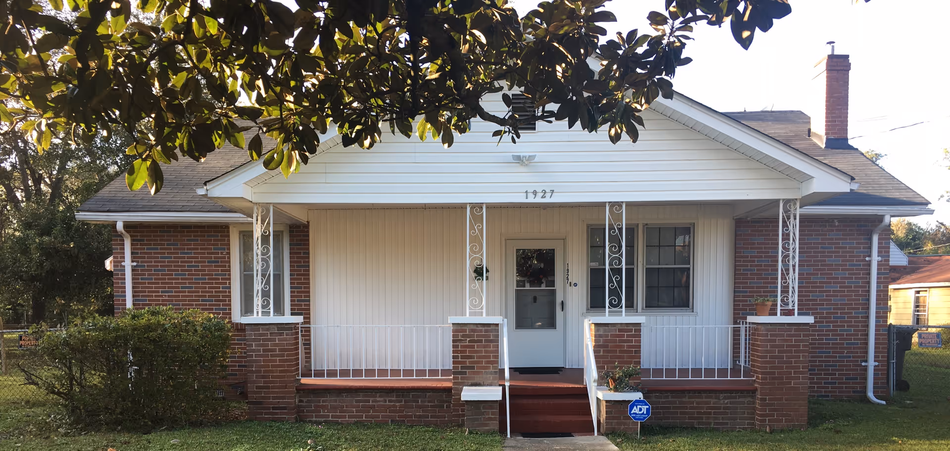Front exterior view of a single-story brick house with a white porch and railing. The house number 1927 is displayed above the porch. There is a small set of stairs leading to the front door, and a blue ADT security sign is placed in the front yard. Trees and bushes surround the house.
