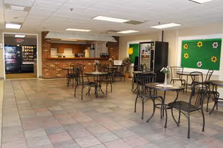 A spacious dining area with several black metal tables and chairs arranged on a tiled floor. The room features a brick counter with a kitchen area behind it, including a refrigerator and vending machines. The walls are decorated with green bulletin boards displaying colorful flower cutouts.
