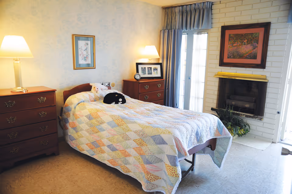 A cozy bedroom with a single bed covered in a colorful patchwork quilt. Two stuffed animals, a white bear and a black cat, are placed on the bed. There are two wooden dressers with brass handles, each topped with a lamp. A framed picture hangs on the wall above the bed, and another framed artwork is above a fireplace with a yellow mantel. Blue curtains cover a glass door next to the fireplace, and a small plant is placed near the fireplace.