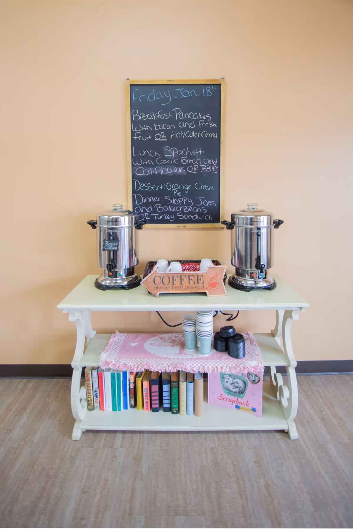 A coffee station with two large metal coffee urns on a white table. Between the urns is a wooden tray labeled 'COFFEE always fresh brewed' holding white coffee cups. Below the table are disposable coffee cups and lids on a pink cloth, and a lower shelf with a row of books and a scrapbook. Behind the table is a chalkboard menu listing meals for Friday, Jan. 18, including breakfast, lunch, dessert, and dinner options.