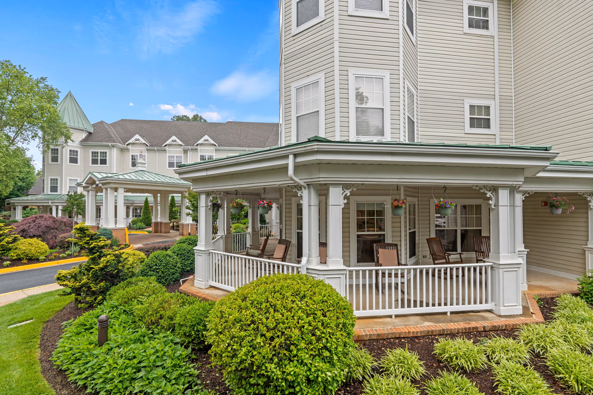 Exterior view of a senior living facility with a covered porch featuring several rocking chairs and hanging flower pots. The building has light-colored siding and multiple windows. Well-maintained landscaping with green bushes and plants surrounds the porch area under a partly cloudy blue sky.