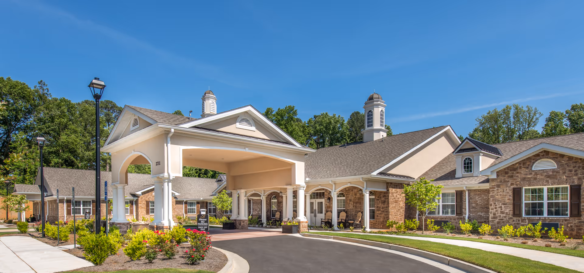 Exterior view of Benton House of Decatur, showing a single-story building with a covered entrance supported by white columns, surrounded by landscaped gardens and trees under a clear blue sky.