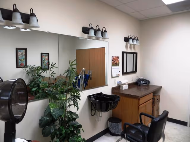 Interior view of a salon or grooming area in a senior living facility. The room features a large wall mirror with three light fixtures above it, a black hair washing sink mounted on the wall, a salon chair, a wooden counter with a radio and tissue box, and a potted plant in the corner. The walls are light-colored, and there is a calendar and framed pictures on the wall.