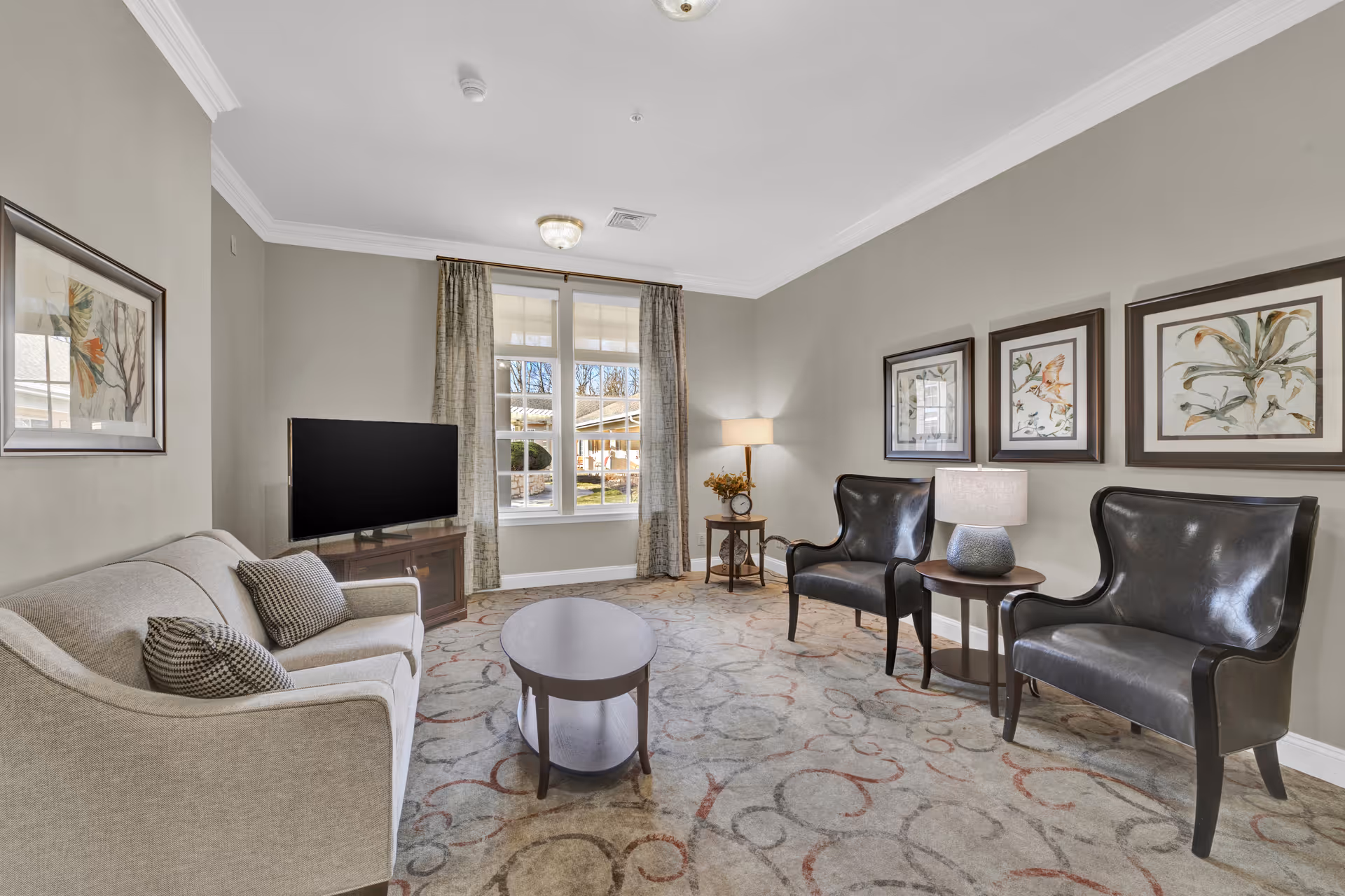 A cozy living room with a beige sofa on the left, two black leather armchairs on the right, a wooden coffee table in the center, and a TV on a wooden stand near a window with curtains. The walls are painted light gray and decorated with framed botanical artwork. A floor lamp and a table lamp provide warm lighting.