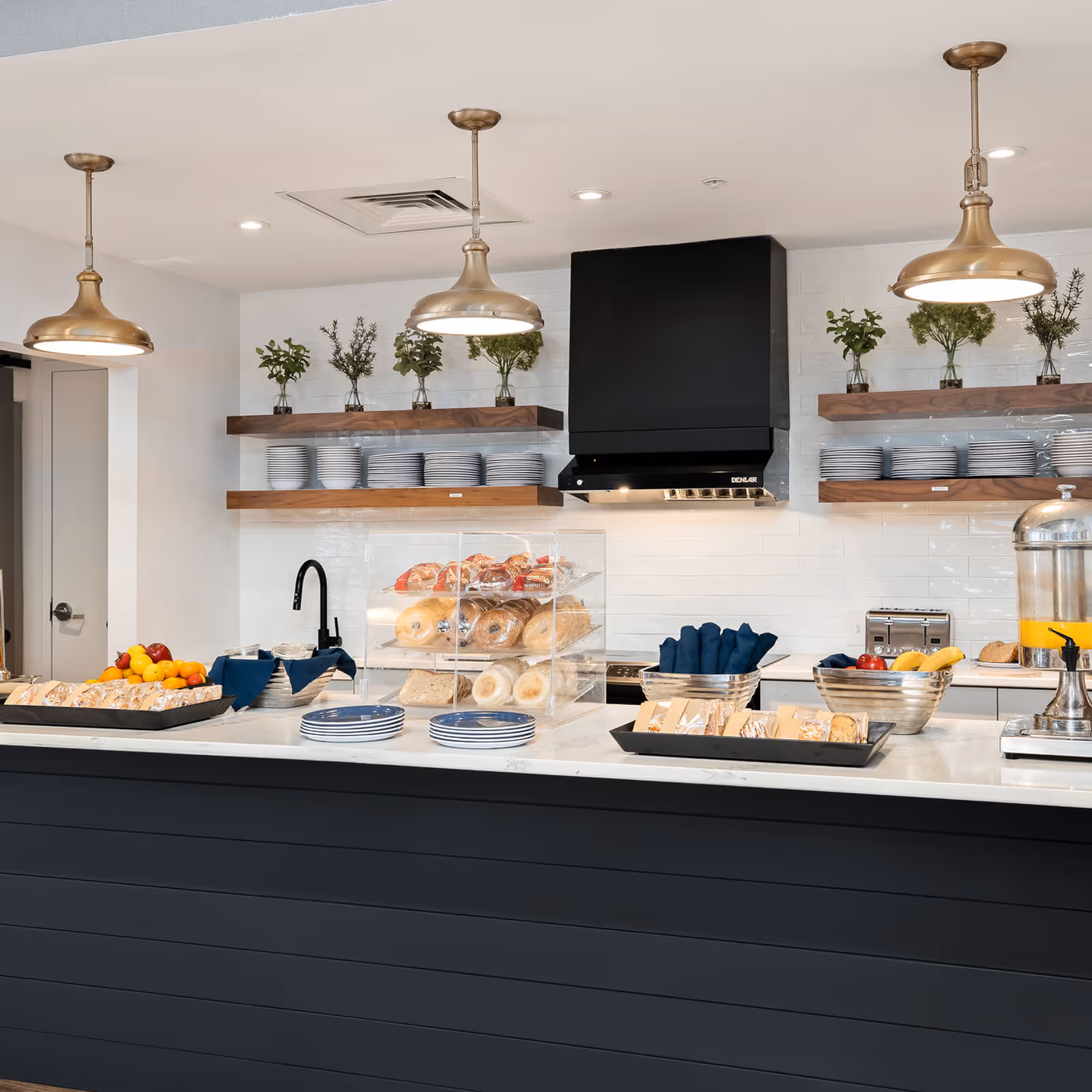 Modern kitchen area with a black and white color scheme featuring a countertop with plates, trays of sandwiches and fruits, a bread display case, and a beverage dispenser. Above the counter are three brass pendant lights, wooden shelves with plates and bowls, and small plants in glass vases. A black range hood is mounted on a white tiled wall.