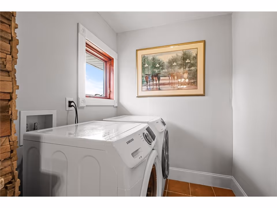 Laundry room with a white Samsung washing machine and dryer side by side, a small window with a red frame letting in natural light, a framed painting of horses on the wall, and a section of stacked stone wall on the left.