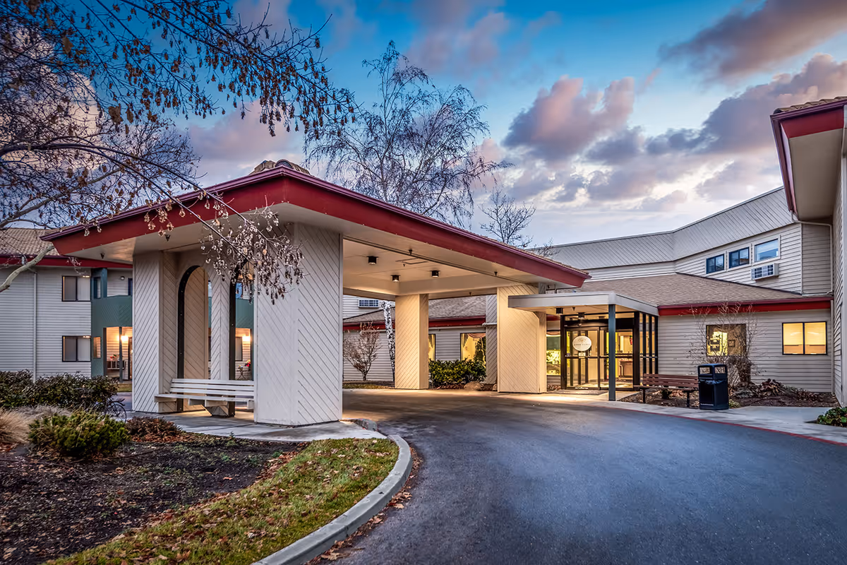 Exterior view of Arbor Village At Hillcrest senior living facility entrance during dusk, showing a covered drop-off area with benches, surrounding trees, and a partly cloudy sky.