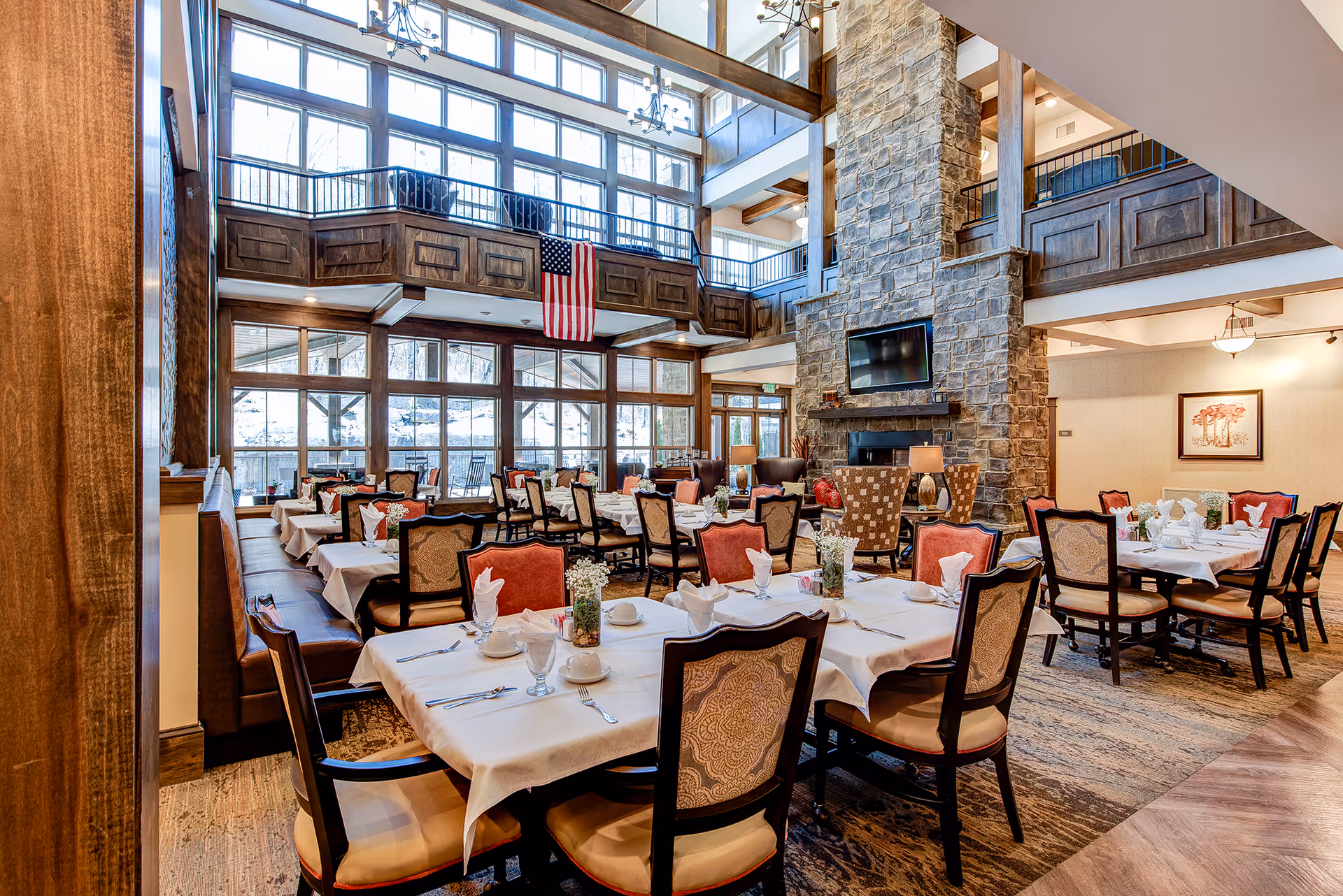 A spacious dining room with multiple tables covered in white tablecloths, set with napkins, cups, and utensils. The room features large windows letting in natural light, a stone fireplace with a mounted TV above it, and a high ceiling with wooden beams. There is an American flag hanging from the balcony railing above.