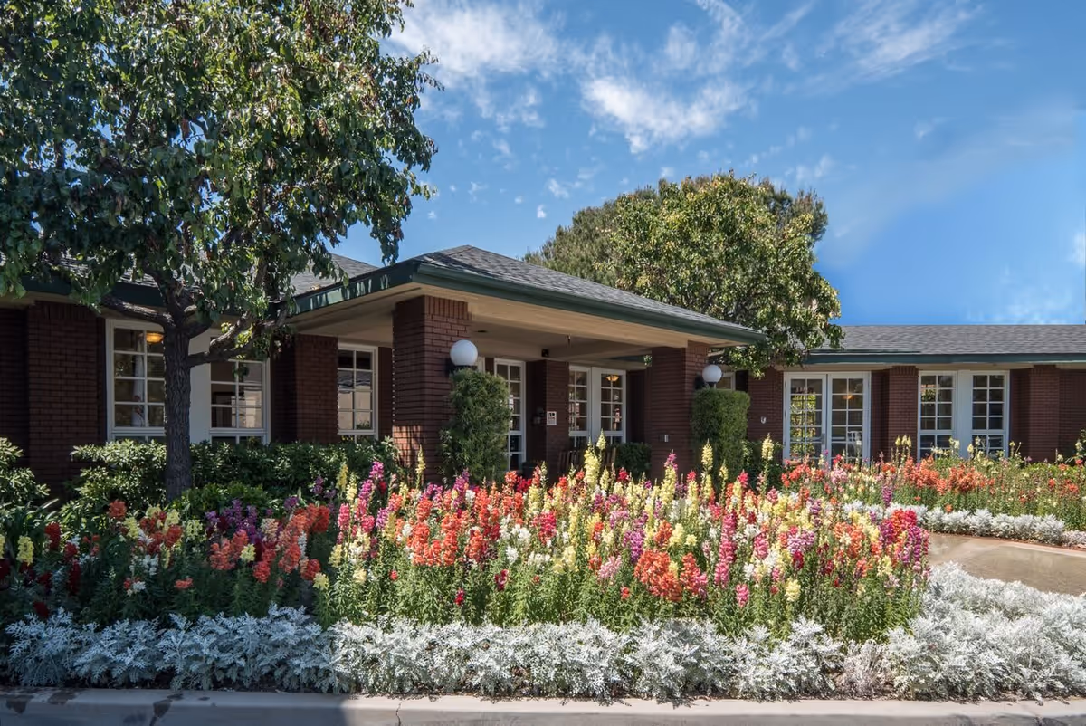 Exterior view of a single-story brick building with large windows, surrounded by a colorful garden filled with various blooming flowers and green trees under a blue sky.
