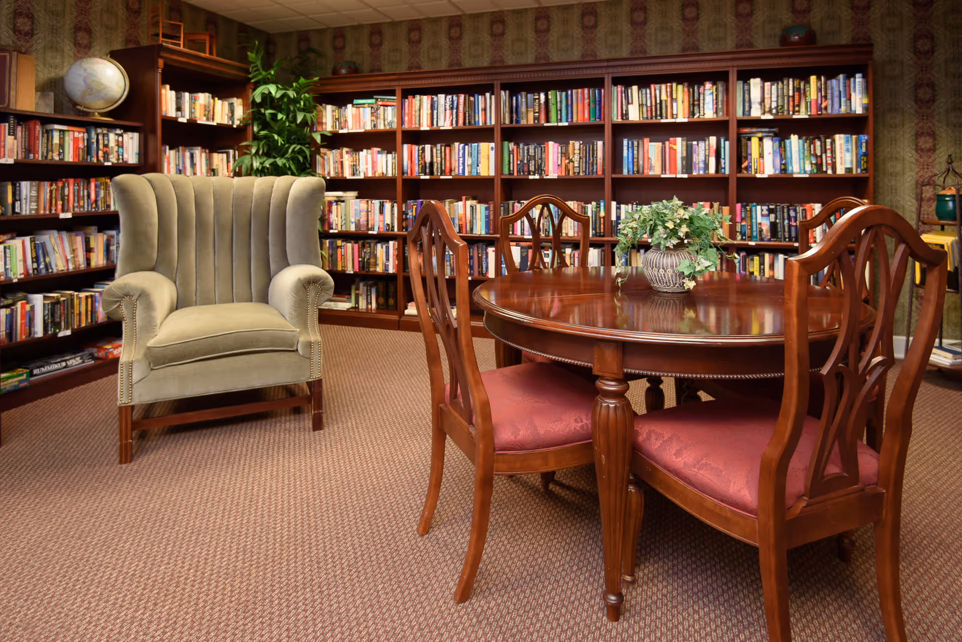 A cozy library room with wooden bookshelves filled with books along the walls, a green upholstered armchair, and a round wooden table with four chairs that have red cushioned seats. A small potted plant is placed on the table.