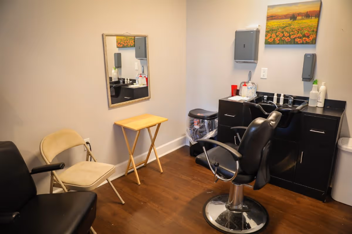 A small salon or grooming room with a black salon chair in front of a black cabinet with a sink. There is a beige folding chair and a small wooden table against the wall. A mirror hangs on the wall above the table, and a colorful painting of a field of flowers is mounted above the sink. The floor is wooden, and the walls are light-colored.