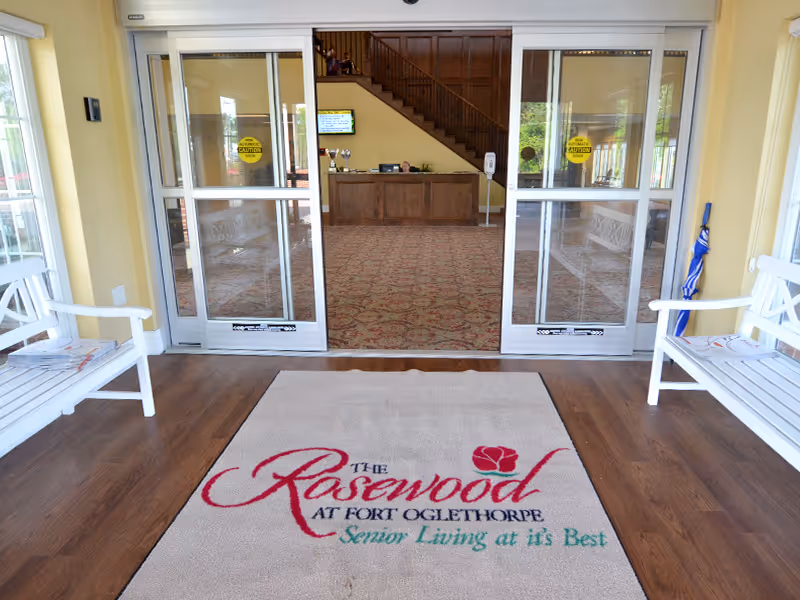 Entrance area of The Rosewood at Fort Oglethorpe senior living facility showing a beige floor mat with the facility's name and slogan, two white benches on either side, and glass automatic sliding doors leading into a lobby with a wooden reception desk and a staircase in the background.