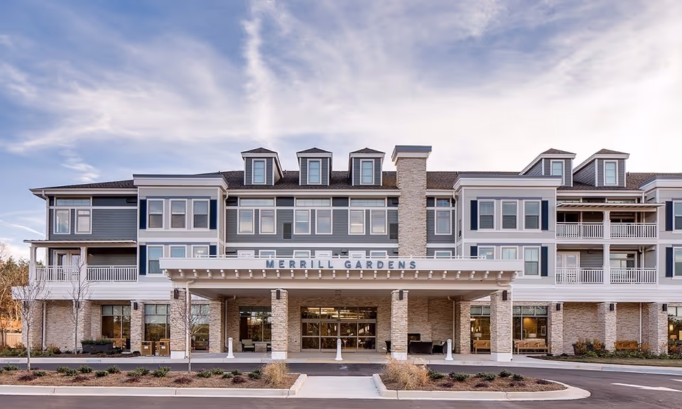 Front exterior view of Merrill Gardens at Woodstock, a multi-story senior living facility with a covered entrance, large windows, balconies, and a stone facade under a partly cloudy sky.