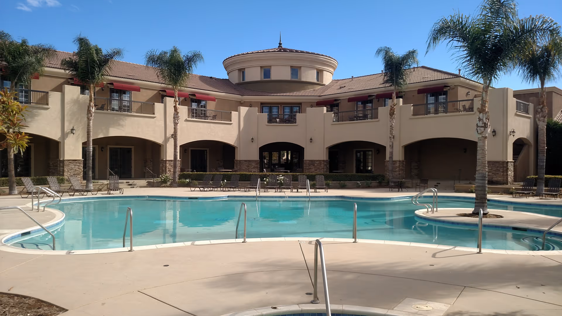 Outdoor swimming pool in front of a two-story Mediterranean-style senior living building with palm trees and lounge chairs.