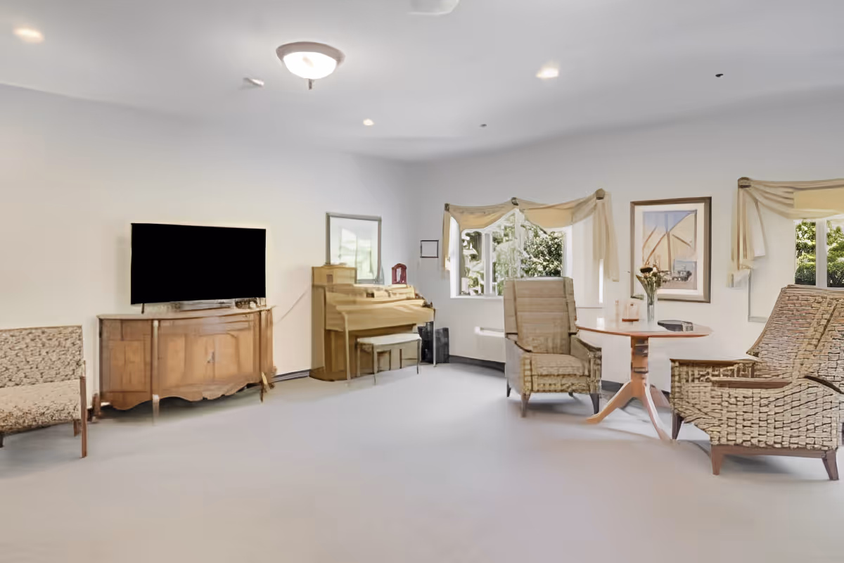 A bright living room with a flat-screen TV on a wooden cabinet, a piano with a bench, two armchairs around a small round wooden table, and windows with light-colored curtains letting in natural light. The walls are white and decorated with framed artwork.