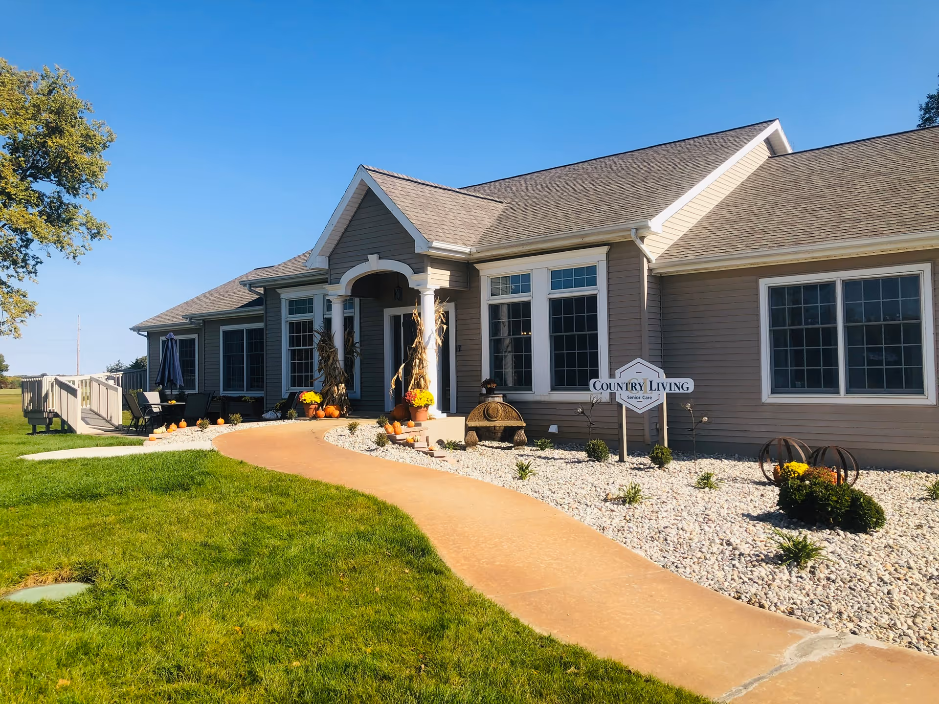 Exterior view of a single-story senior care facility building with a beige facade, multiple windows, and a peaked roof. A curved concrete walkway leads to the entrance, which is decorated with pumpkins, flowers, and cornstalks. There is a sign near the entrance that reads 'Country Living Senior Care'. The surrounding area includes green grass, white gravel landscaping, and a clear blue sky.