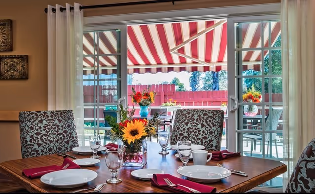 Dining table set with plates, glasses, and a floral centerpiece in front of sliding glass doors leading to a patio with a red-and-white striped awning.