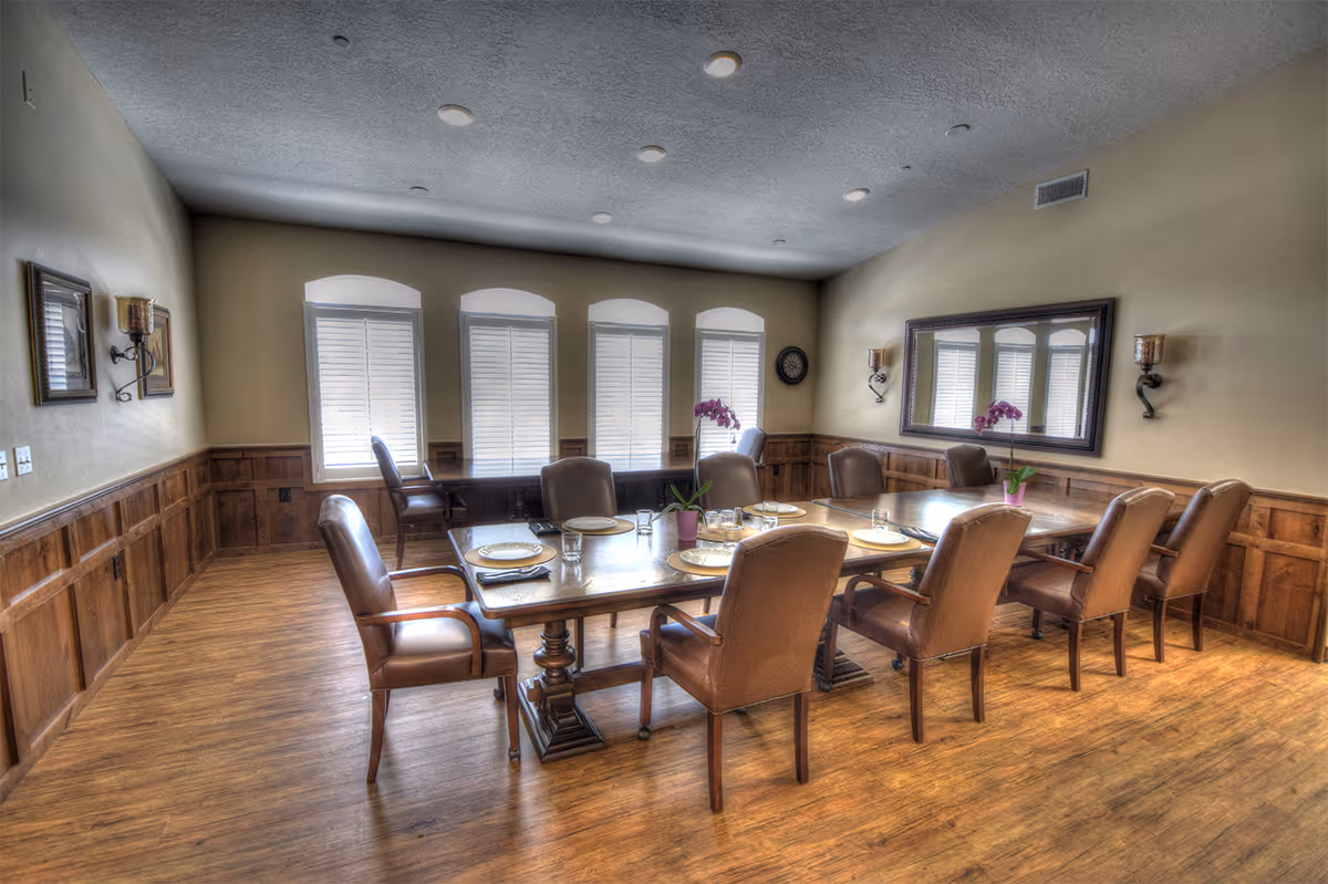 A formal dining room with a long wooden table set for a meal, upholstered chairs, wood-paneled walls and shuttered windows.