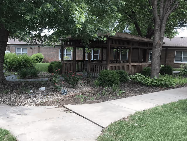 Shaded outdoor courtyard with a wooden gazebo, trees, landscaping, and a brick building in the background.