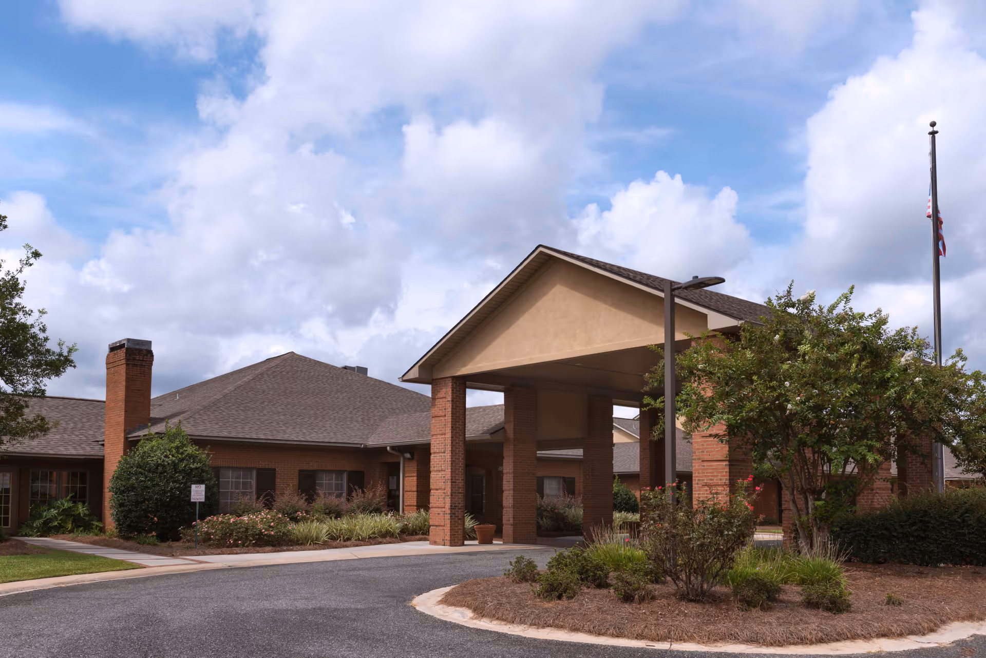 Exterior view of Magnolia Manor in Moultrie showing a single-story brick building with a covered entrance supported by brick columns, surrounded by landscaped bushes and trees under a partly cloudy sky.