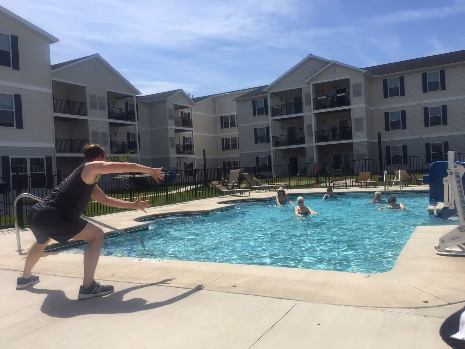 People swimming in an outdoor pool while a person on the deck gestures, with multi-story apartment buildings behind them.