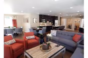 A spacious and well-lit living room area featuring a gray sofa with red and patterned cushions, two red armchairs, a wooden coffee table with a plant centerpiece, and a dining area with a table and chairs in the background. The room has recessed lighting and a modern kitchen with dark cabinetry and stainless steel appliances visible behind the dining area.