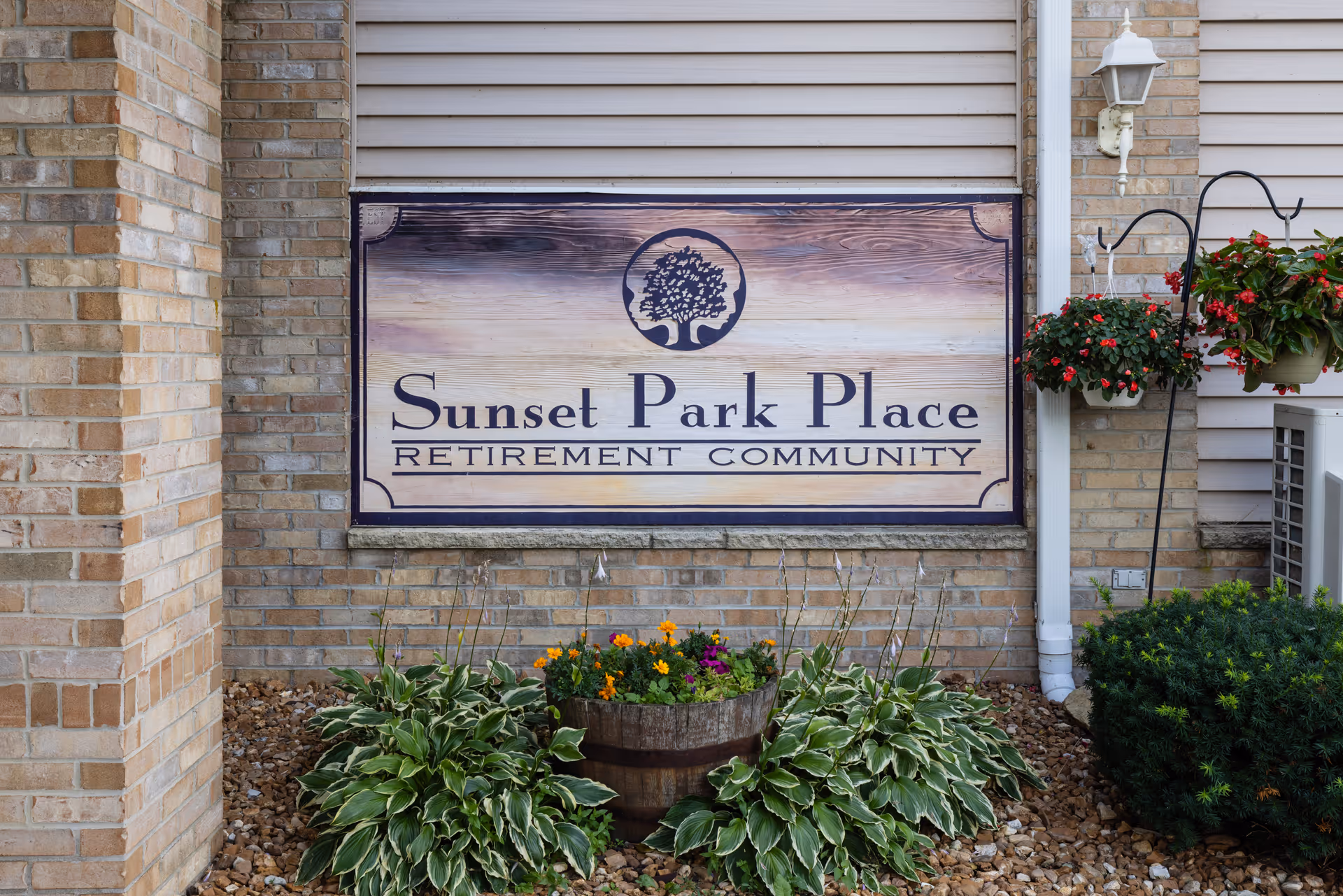A wooden sign mounted on a brick wall that reads 'Sunset Park Place Retirement Community' with a tree logo above the text. The sign is surrounded by plants and flowers in front of the wall, with a hanging flower pot on the right side.