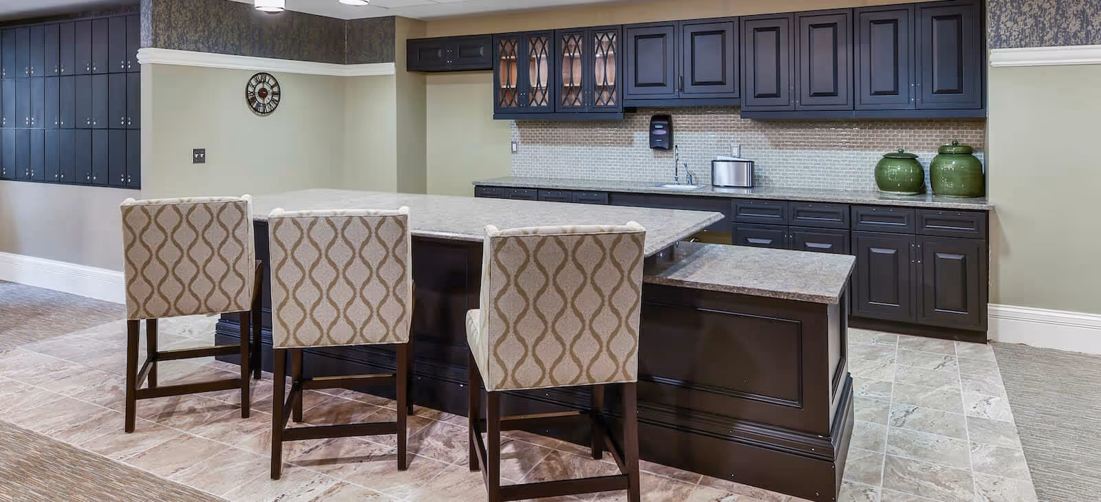 Interior view of a kitchen area in a senior living facility with a large island countertop surrounded by three patterned upholstered chairs. The kitchen features dark cabinetry, a tiled backsplash, and various kitchen appliances and decor items on the counters.
