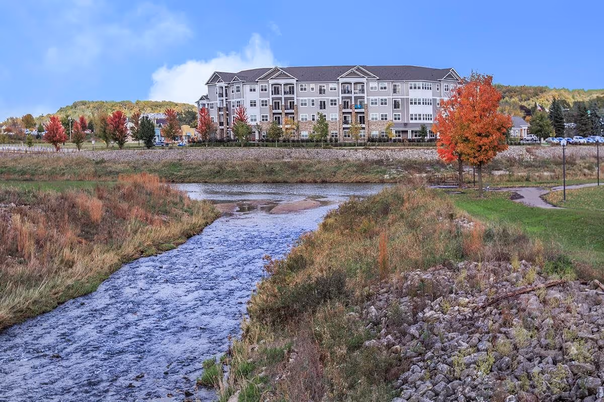 A large multi-story senior living facility building named AVIVA River Bend is visible in the background, surrounded by trees with autumn foliage. In the foreground, there is a flowing river bordered by grassy and rocky banks under a clear blue sky.