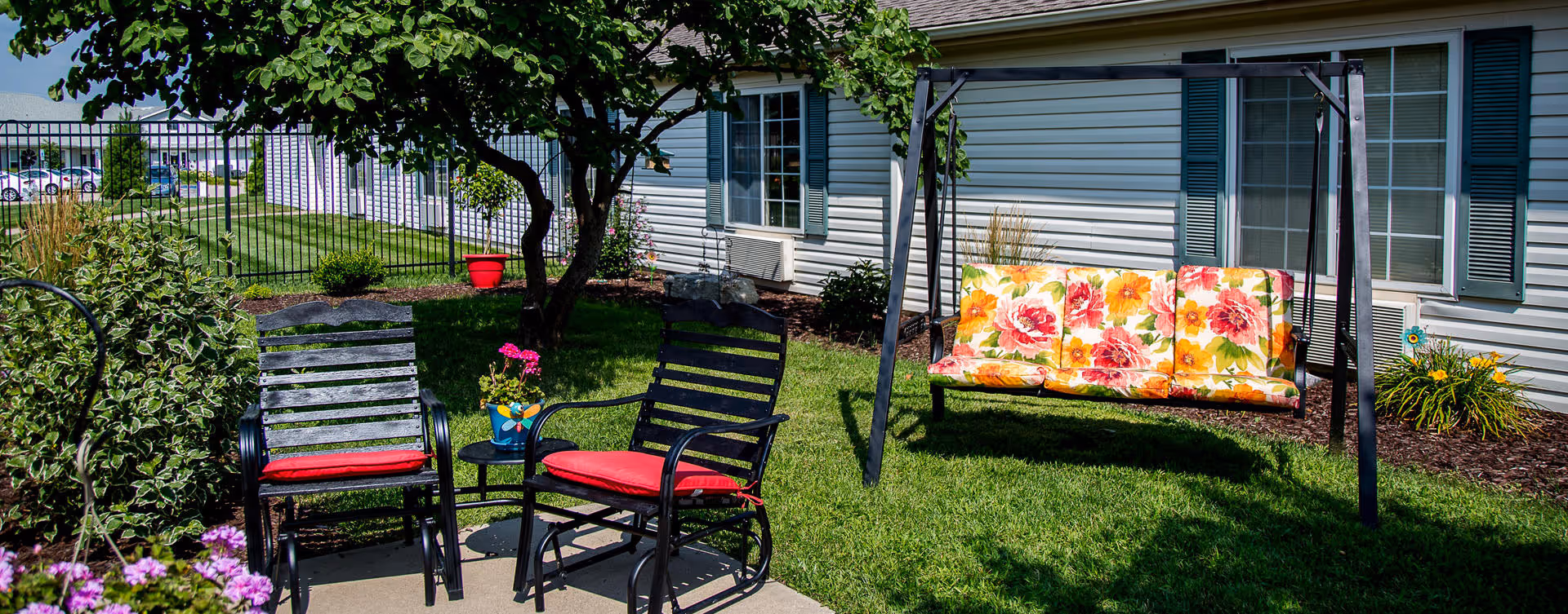 Outdoor seating area with two black metal chairs with red cushions, a small round table with a potted pink flower, and a floral cushioned swing set on a green lawn next to a building with white siding and windows.