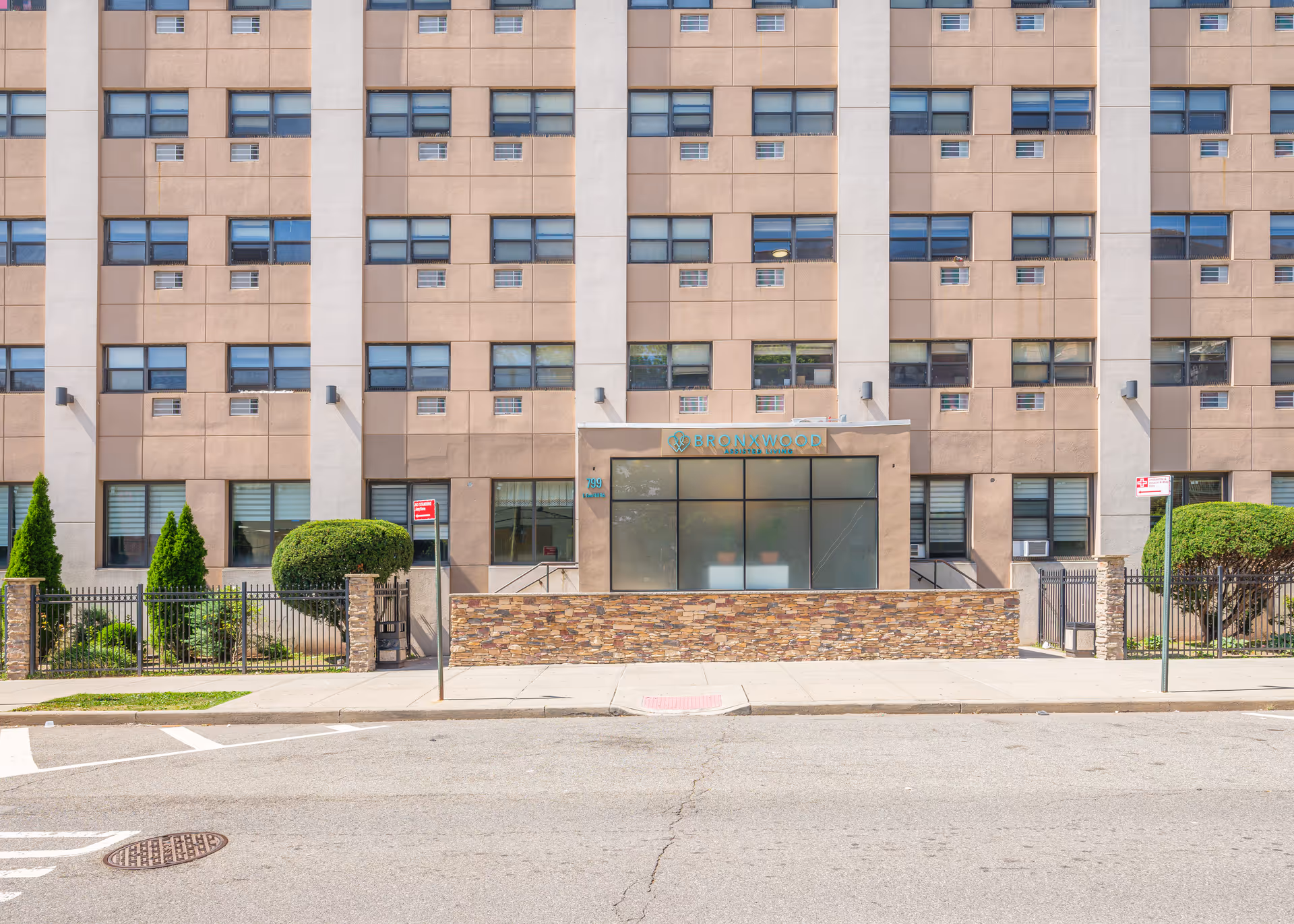 Front exterior view of Bronxwood Assisted Living building with multiple windows, a stone wall, and a sidewalk in front.