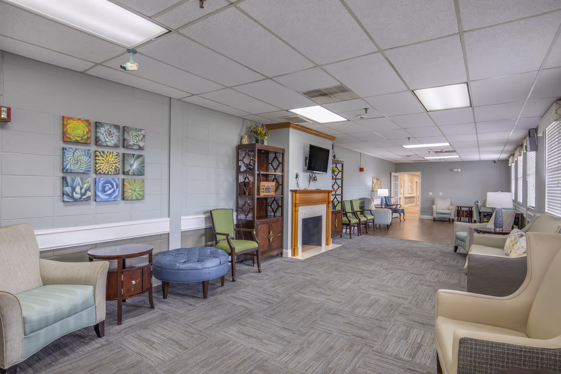 Spacious common lounge with armchairs, ottoman, fireplace and wall-mounted TV in a senior living facility.