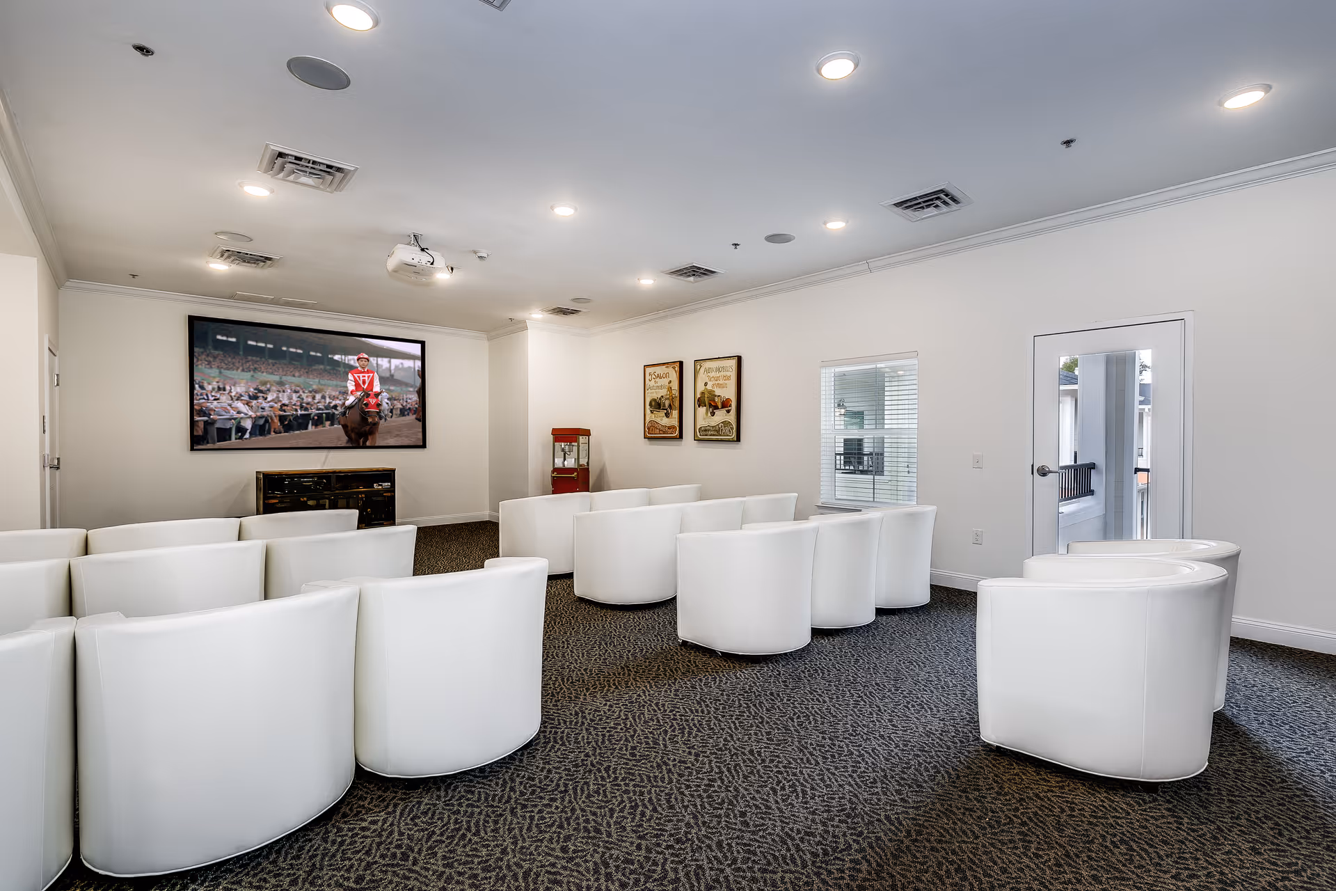 A small screening room with rows of white curved chairs facing a large wall-mounted TV showing a horse race, with a popcorn machine and framed posters on the back wall.