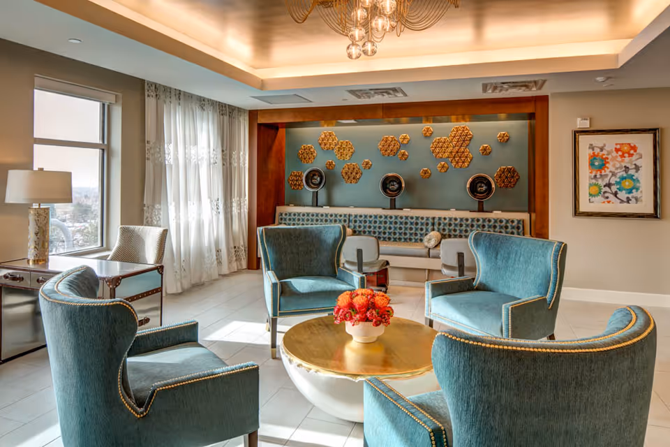 A bright and elegant living room area with four teal upholstered armchairs arranged around a round coffee table with a flower arrangement. The room features a large window with sheer curtains, a side table with a lamp, a decorative wall with geometric wooden art pieces, and a framed colorful artwork on the wall.