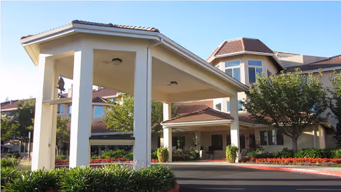 Porte-cochere entrance of a multi-story retirement community building with manicured landscaping and a clear blue sky.