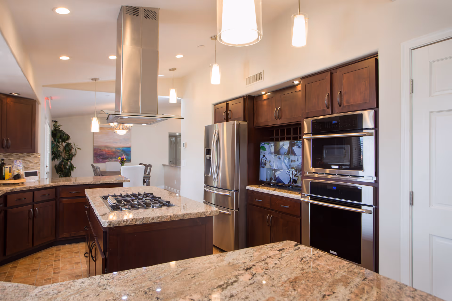 Modern kitchen with granite countertops, dark wooden cabinets, a stainless steel refrigerator, double wall ovens, and a stovetop on a central island with a stainless steel range hood above. Pendant lights hang from the ceiling, and a dining area with a table and chairs is visible in the background.