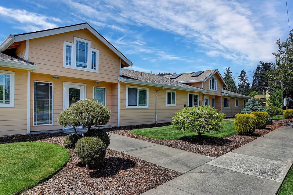 Exterior view of a single-story residential care facility building with beige siding, multiple windows, and a small landscaped garden with trimmed bushes and green grass under a partly cloudy blue sky.