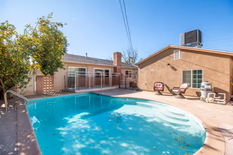 Outdoor courtyard with a kidney-shaped swimming pool next to single-story buildings, lounge chairs, and a tree under a clear sky.