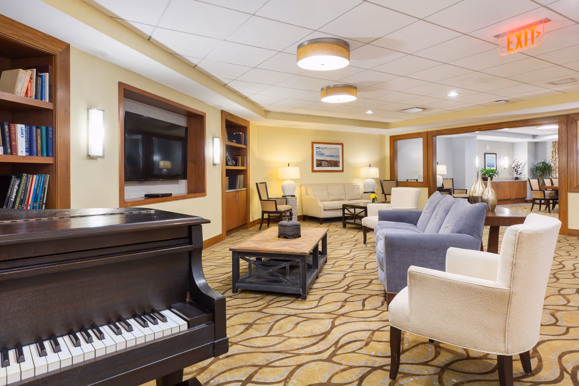 A cozy senior living facility common area featuring a dark wooden piano in the foreground, a large flat-screen TV mounted on the wall, bookshelves, and comfortable seating including armchairs and sofas arranged around a wooden coffee table. The room is well-lit with ceiling lights and table lamps, and has a patterned carpet and light yellow walls.